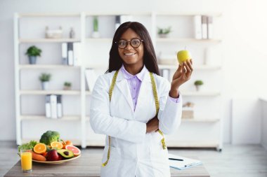 Bir beslenme uzmanı parlak bir mutfakta elinde yeşil bir elmayla durup gülümsüyor. Taze meyve ve sebzeler yakınlardaki bir masada sergilenir. Bu sahne sağlıklı beslenmeyi teşvik ediyor..