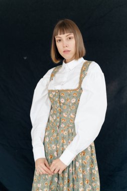 Eski moda elbiseli genç bir kadın stüdyoda karanlık arka planda poz veriyor.