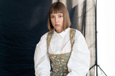 Eski moda elbiseli genç bir kadın stüdyoda karanlık arka planda poz veriyor.