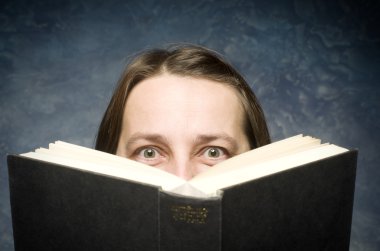 Mavi arka plan üzerinde kitap arkasına saklanmış mutlu olgun kadın.