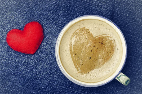 Kopje koffie met schuim in de vorm van hart op blue jeans, denim achtergrond cappuccino. Rood hart handgemaakte van vilt, dag van de Valentijnskaart concept. — Stockfoto