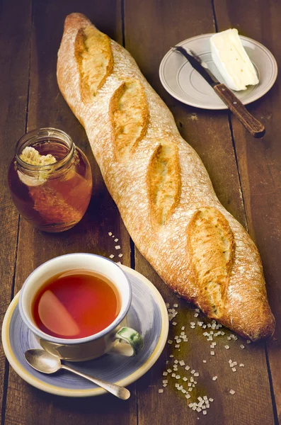 Hemlagad färsk baguette, plattan med ost, burk naturlig honung och kopp te på träbord. — Stockfoto