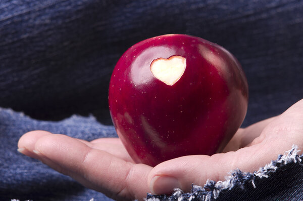 Fresh red apple with a heart shaped cut-out in woman hand on denim, jeans background. GMO free genetically modified organisms. Mothers day
