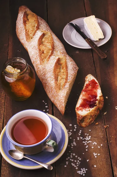 Bit av hemlagad färsk baguette med sylt, plattan med ost, burk naturlig honung och kopp te på träbord. — Stockfoto