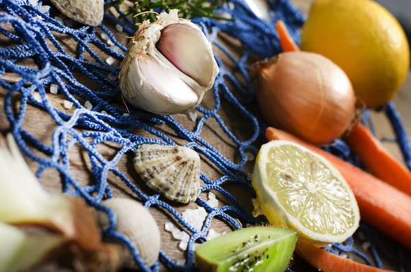 Fondo de cocina. Ajo, cebolla, zanahoria, limón sobre un fondo de madera. Estilo marítimo, una red de pesca azul y conchas . — Foto de Stock