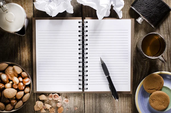 Ahşap masa boş sayfaları ile açık bir defter. Fincan çay, hip flask, fındık ve bisküvi aperatif.