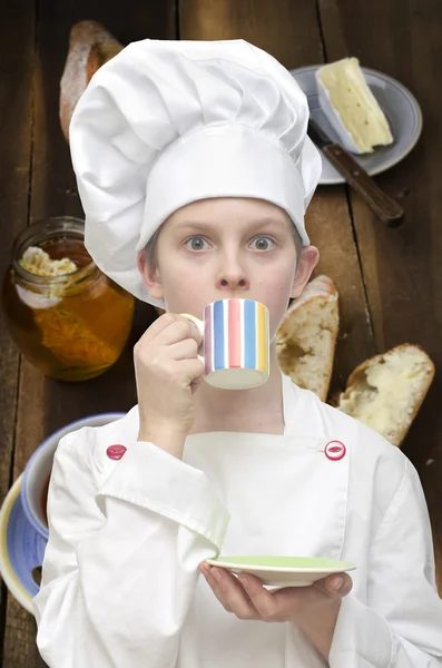 Young boy dressed as chef drinking tea on food background. — Stock Photo, Image