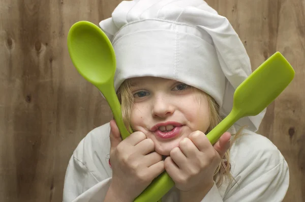 Young girl dressed as chef with green silicone spoons on wooden background. — Stock Photo, Image