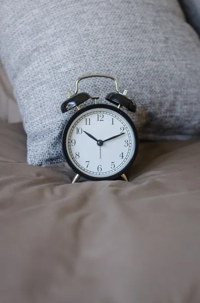 Black alarm clock on brown bed with grey pillow — Stock Photo, Image