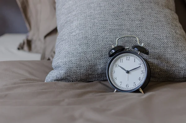 Μαύρο ρολόι συναγερμών σε καφέ κρεβάτι με μαξιλάρι γκρι — Φωτογραφία Αρχείου