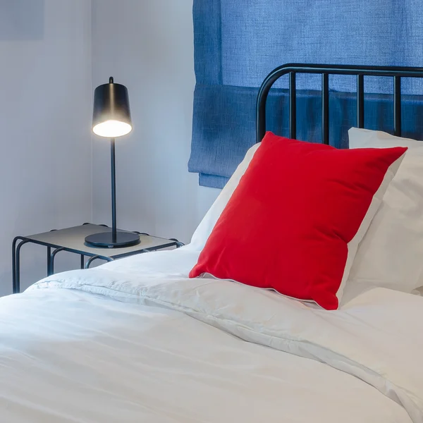 Cuscino rosso su letto bianco con tenda blu e lampada nera su tabl — Foto Stock