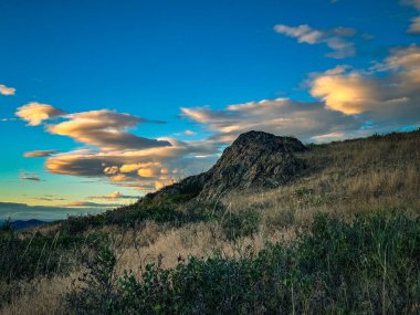 Vahşi çimenli bir yamaçtan yükselen kayalık bir tepenin görkemli manzarası. Dramatik günbatımı bulutları, uzak bir dağ sırası ve göl üzerinde mavi gökyüzünü doldurur. Açık havanın özünü yakalar..