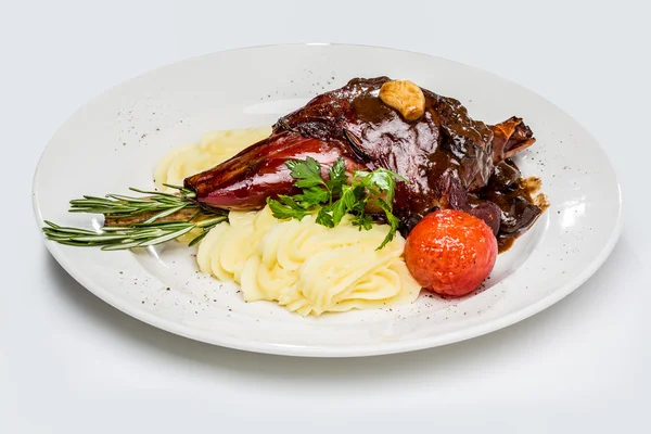 Caña de cordero asado, marinado en vino tinto con ciruelas pasas y ajo. con puré de papas y tomates al horno. Sobre un fondo blanco y un plato blanco —  Fotos de Stock