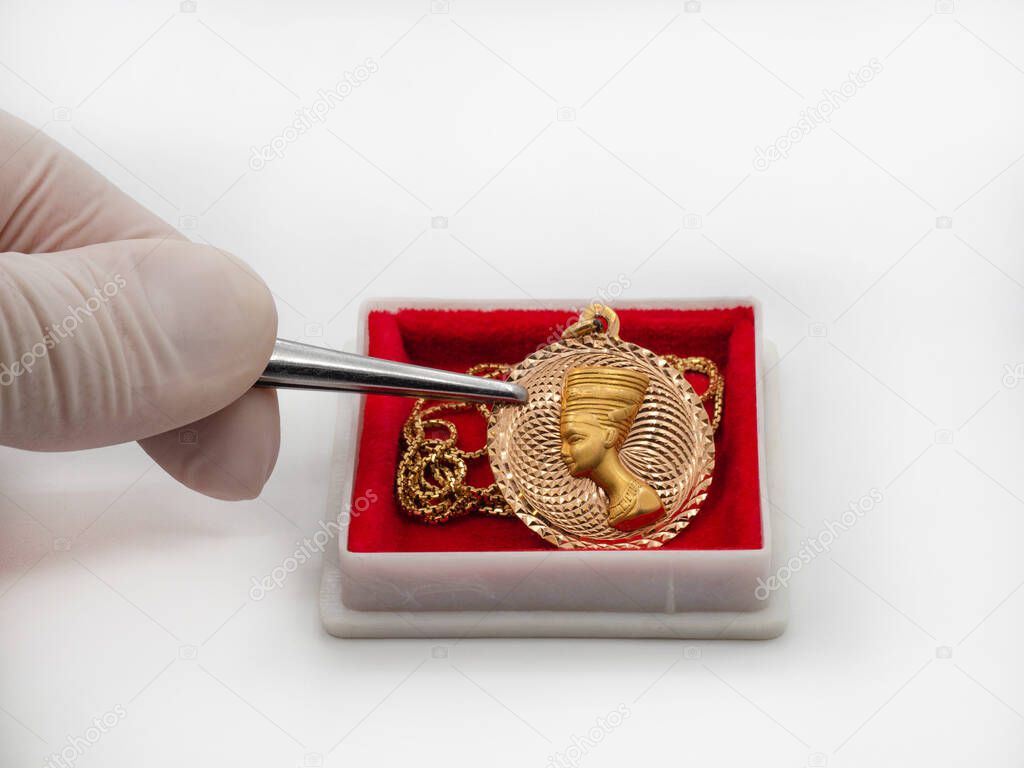A gloved hand with tweezers holds a gold female pendant in the form of a portrait of Cleopatra with a chain on a red cushion in a jewelry case. The concept of jewelry valuation, sale, auction.