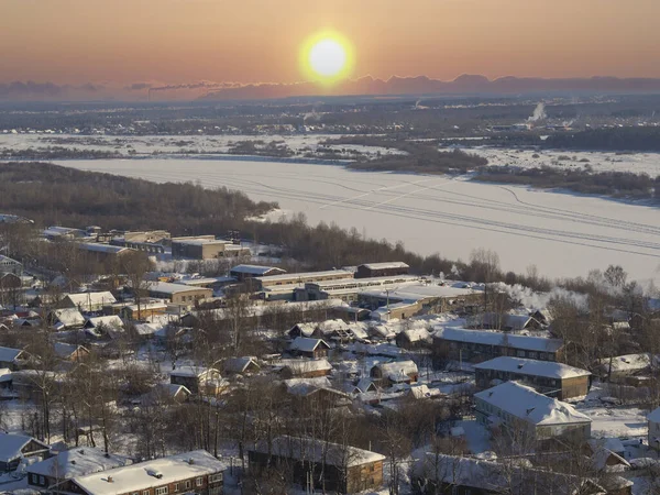 A suburb, village, or industrial quarter of a winter city (panorama from skyscraper). Russian hinterland, view of the frozen ice of the Vyatka River. Sunrise, cold dawn over the wooden houses.