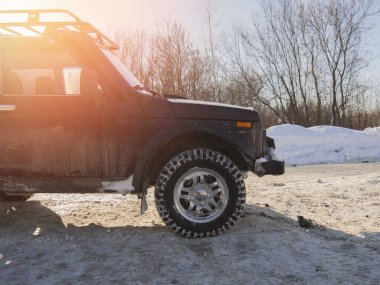 Bahar güneşinde toprak bir yola parketmiş bordo bir Rus SUV. Karda dört çeker bir arabanın 32 inçlik tekerlekleri. Sefer sepeti, raylar ve bariyerler arabada..