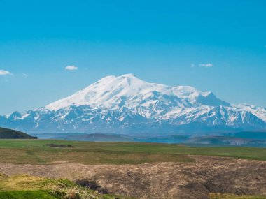 Gumbashi geçidinin sarı yay alp çayırlarında yeşil çimenler. Kar mavi gökyüzüne karşı ufukta Elbrus Dağı 'nı kapladı. Kafkas Dağları 'nın dağlık, tepelik yaz manzarası.