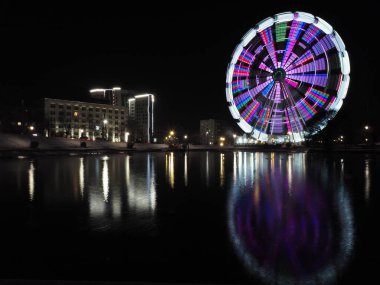 Ferris wheel at long exposure at night. Live composite of the attraction in the park in winter. Reflections of light and color in the lake water.