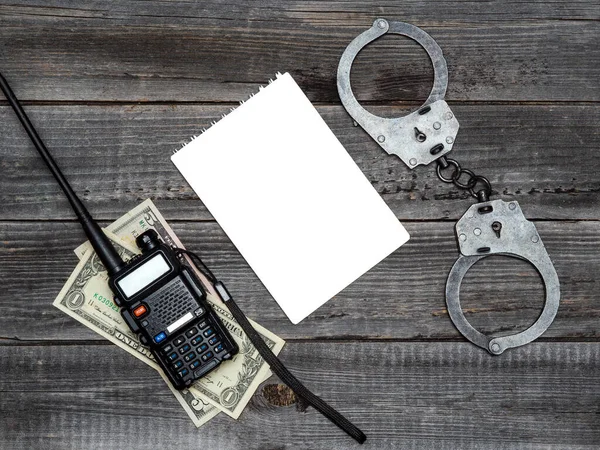 A portable walkie-talkie with dollar bills and handcuffs on a wooden table background. Copy space white notepad. The concept of police corruption, black market, money crime.