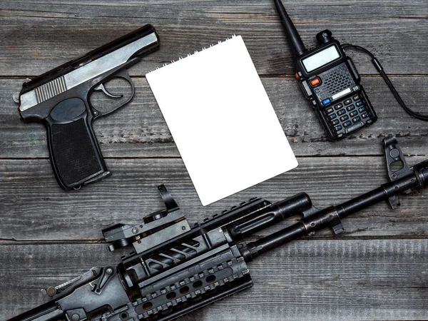 一把战斗自动步枪 一把警用手枪和一个手提式收音机 放在一张桌子的木制背景上 桌上有一张白色的记事本 平铺气垫布局 — 图库照片