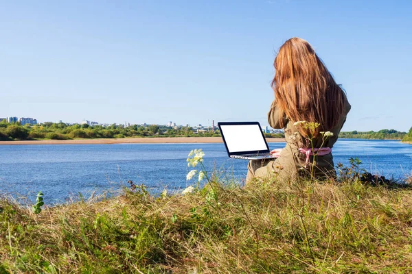 A girl with a laptop is sitting on the river bank in a park outside the city. The concept of freelance, remote work or training. An independent female businessman works in nature while traveling.