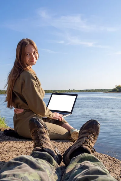A girl with a laptop is sitting on the river bank in a park outside the city. The concept of freelancing, remote work or training. A young couple of lovers are resting in a city park.
