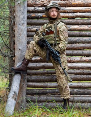 Askeri üniformalı bir kız elinde otomatik bir tüfek tutuyor. Kurşungeçirmez yelek giymiş kadın asker ve ormanda tahta duvarın arkasında taktik miğfer. Eğitim kampı, atış poligonu..