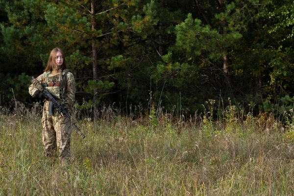 A girl with red hair in camouflage clothing and a bulletproof vest stands in a field against the background of a forest. A military woman holds an automatic rifle in her hands.