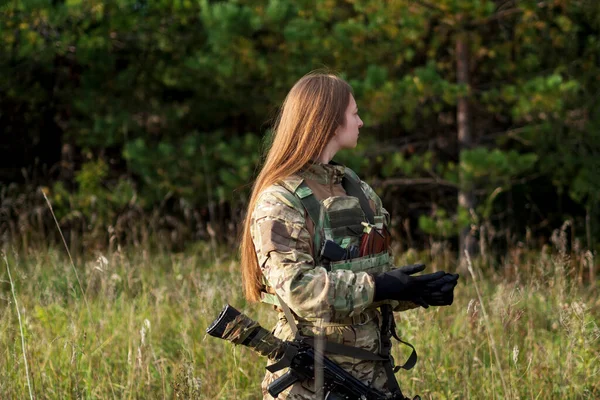 A girl with red hair in camouflage clothing and a bulletproof vest stands in a field against the background of a forest. A military woman holds an automatic rifle in her hands.