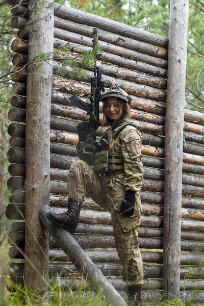 Girl in military uniform holds an automatic rifle in her hands. Woman soldier in bulletproof vest and tactical helmet in the forest against background of wooden wall. Training camp, shooting range.