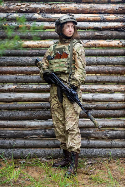 軍の制服を着た少女は手に自動小銃を持っている 防弾と木製の壁を背景に森の中で戦術的なヘルメットの女性兵士 訓練キャンプ射撃場 — ストック写真