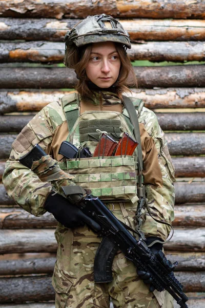 Girl in military uniform holds an automatic rifle in her hands. Woman soldier in bulletproof vest and tactical helmet in the forest against background of wooden wall. Training camp, shooting range.