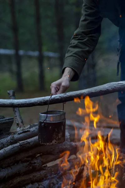 Man hangs up military army pot on fire in the dark. Cooking food in a kettle or tea on a campfire against the background of the forest. Survival and tourism.