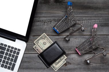 Bilgisayarın arka planında siyah bir cüzdanın yanında gri ahşap bir masanın arkasında dolar ve alışveriş arabası var. Düz yerde, üst tarafta. Kara Cuma günü online alışveriş kavramı.