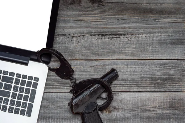 Elleri kelepçeli siyah bir polis tabancasının yanındaki gri ahşap bir masanın arka planına karşı dizüstü bilgisayar. Düz yerde, üst tarafta. İnternet güvenliği ve online suç kavramı.