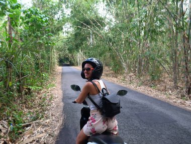 Güvenli kaskı ve güneş gözlükleriyle kiralık motosiklet süren genç, mutlu, beyaz bir kadın Bali adasında güzel bir bambu ormanını gezerken güvenli bir şekilde araba kullanıyor.