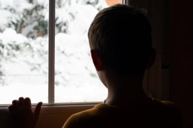 Sırtı ışığa dönük çocuk evinin penceresinden karlı manzaraya bakıyor..