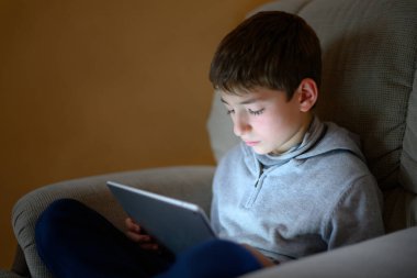 Kanepede oturup tablete bakan ciddi bir çocuk.