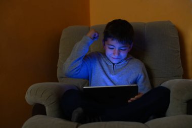 Çocuk kanepede oturmuş tablete bakıyor, yumruğunu kaldırmış ve gülümsüyor.