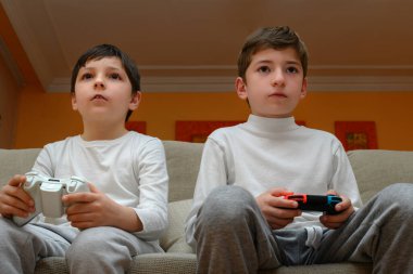 Kanepede oturan iki çocuk bir oyun konsolunun kumandalarını tutuyorlar.