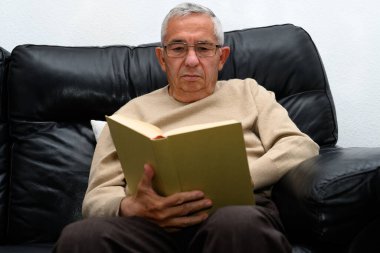 Gözlüklü bir adam evdeki kanepede kitap okuyor.