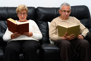 Orta yaşlı erkek ve kadın, her biri kitap okuyor. Evlerinde bir kanepede oturuyorlar..