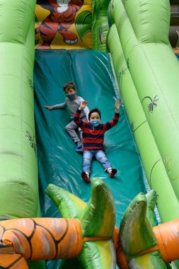 Yüz maskeli iki çocuk şişme bir kaydırağın altına girerken eğleniyor.