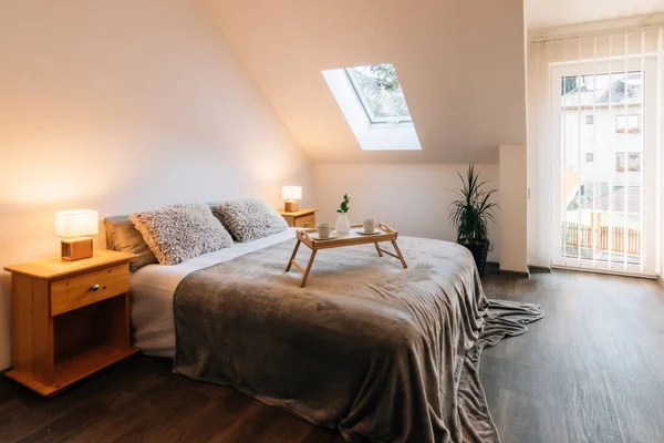 Small attic bedroom in a family house with morning breakfast service on the bed. The bed also has a fluffy blanket and two fluffy pillows, on the sides there are bedside tables with lit lamps.