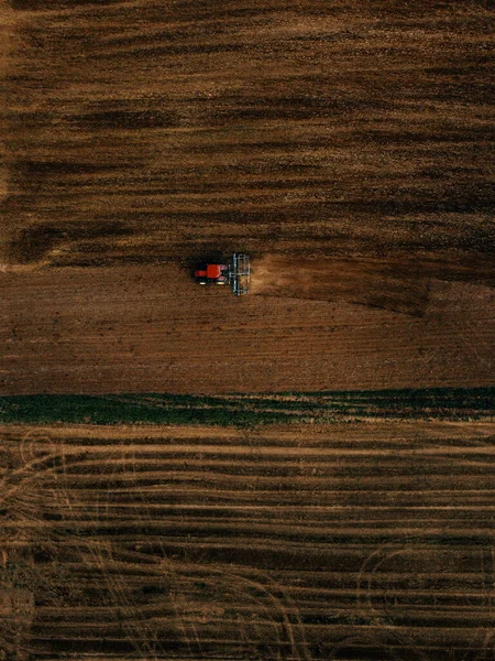 Aerial view of field in which tractor plows.Blue tillage equipment and red tractor are clearly visible on dark brown background of earth with drawings from tillage and wheels and green grassy line.