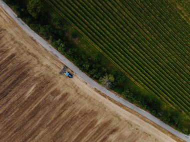 Aerial view of field in which tractor plows.Blue tillage equipment and blue tractor are clearly visible on  brown background of earth.Road divides scape in two halves with green color in other one.