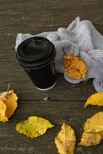 プラスチック製の蓋付きの黒い紙のコーヒーカップは 暖かい柔らかい灰色のスカーフと黄色の葉の横にある木製の屋外テーブルに座っています — ストック写真