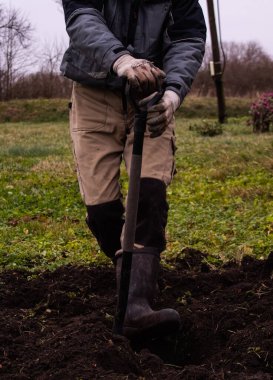 Çalışan üniformalı ve çizmeli bir adam kazmak için toprağı kazıyor. 