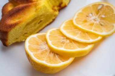 Ön planda dilimlenmiş limon, beyaz bir tabakta fırında limonlu turtanın yanında.