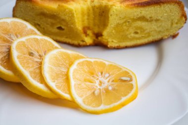 Ön planda dilimlenmiş limon, beyaz bir tabakta fırında limonlu turtanın yanında.